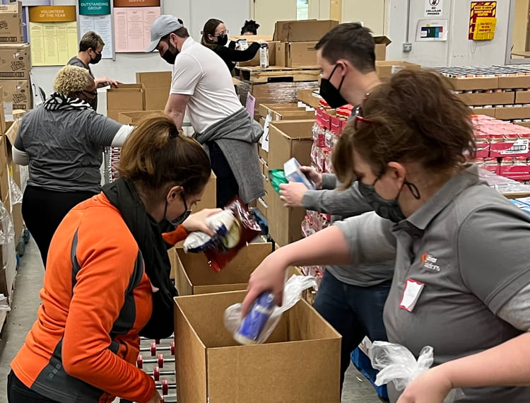 A group of people all wearing masks are working in a warehouse and packing up cardboard boxes with supplies.